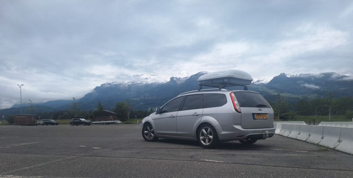 Een zilverkleurige Ford Focus stationwagen met een dakkoffer staat geparkeerd op een asfaltterrein. Op de achtergrond zijn besneeuwde bergtoppen zichtbaar onder een bewolkte lucht. De bergen zijn deels bedekt met groen bos, en er is een licht glooiend landschap te zien. Rechts van de auto staan witte betonnen afscheidingen. Links op de parkeerplaats staan nog enkele andere voertuigen en een lantaarnpaal.
