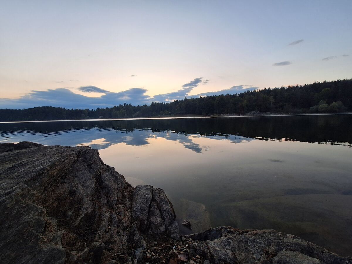 Een rustig meer bij zonsondergang, met de weerspiegeling van de lucht en wolken op het water. Op de voorgrond zijn rotsen en kleine steentjes zichtbaar, en de overkant van het meer is begroeid met bomen.