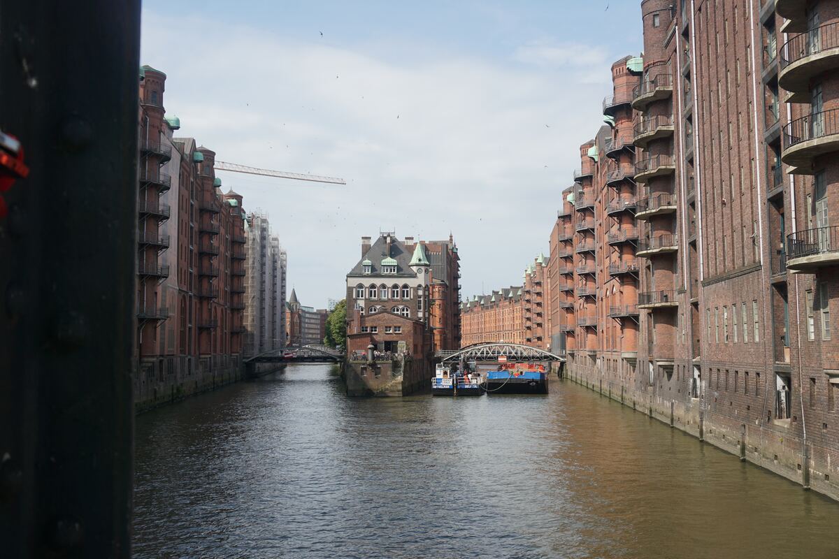 Een foto van een kanaal omringd door historische bakstenen pakhuizen met balkons en groene daken. Op het water liggen enkele boten en in de verte zijn bruggen en een bouwplaats met een kraan zichtbaar. De lucht is licht bewolkt.