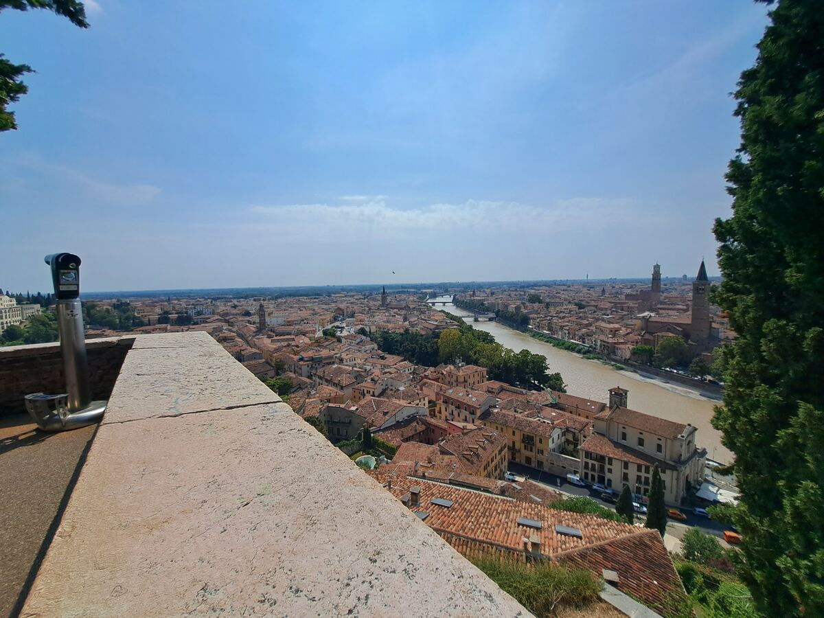 Een panoramisch uitzicht over een historische stad met veel daken van terracotta, een rivier die door de stad stroomt, en diverse kerktorens. Op de voorgrond is een stenen muur met een openbare verrekijker te zien. De lucht is helderblauw met enkele lichte wolken. Aan de rechterkant is een deel van een boom zichtbaar.