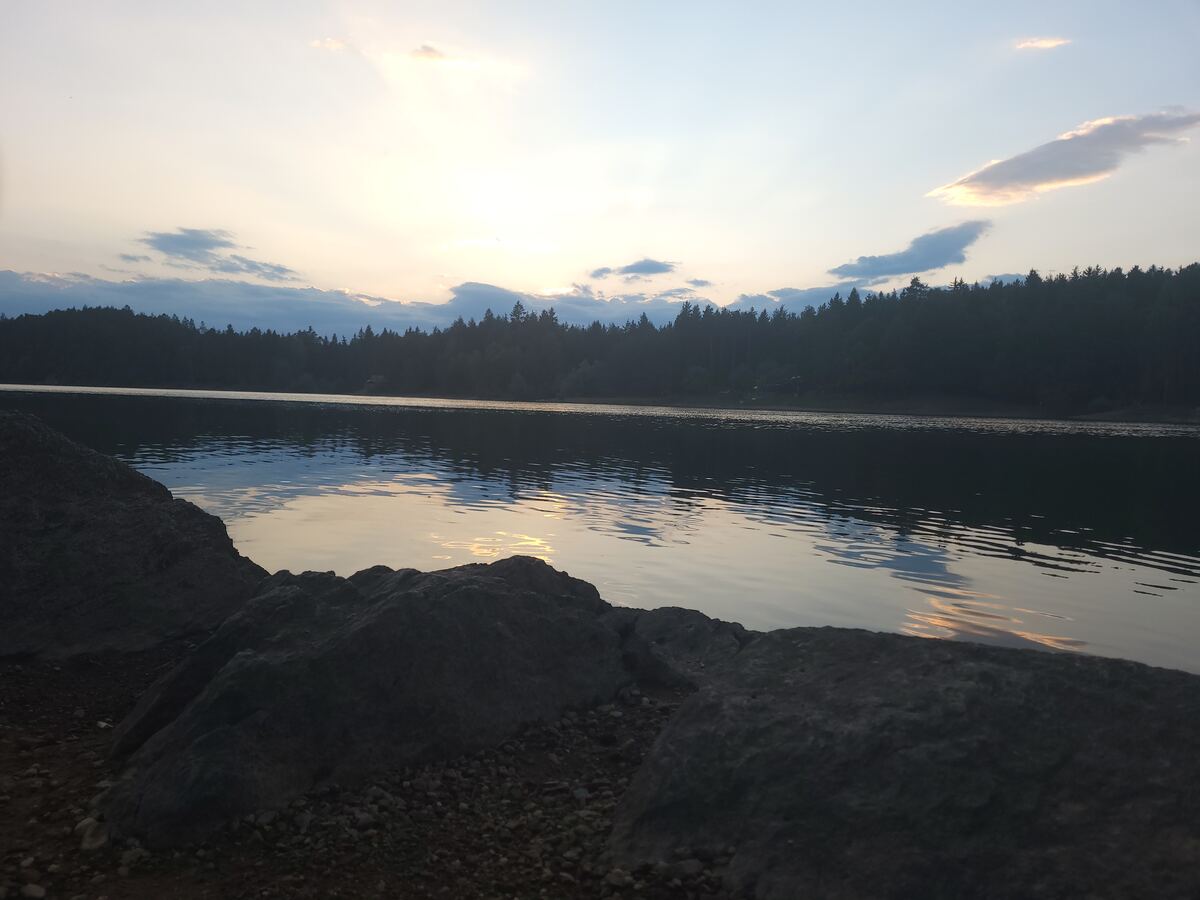 Een meer met donkere, beboste oevers onder een avondlucht met lichte wolken. De zon reflecteert zachtjes op het wateroppervlak. Op de voorgrond liggen grote rotsen en kiezels langs de waterkant.