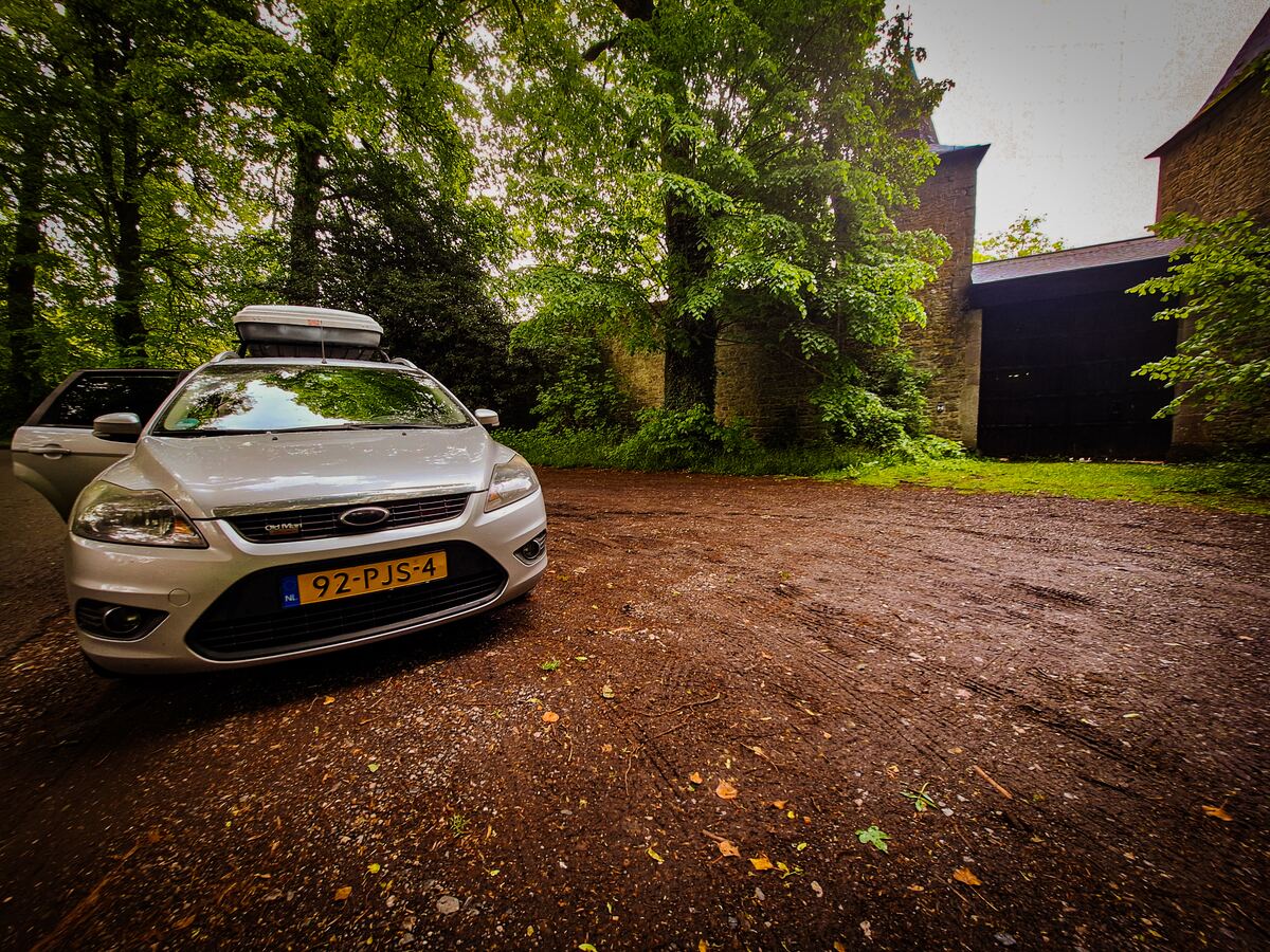 Een zilverkleurige Ford Focus stationwagen met een dakkoffer staat geparkeerd op een onverharde ondergrond. De passagiersdeur aan de voorzijde staat open. Op de achtergrond is een stenen gebouw te zien, mogelijk een kasteel of landhuis, omgeven door veel groen en bomen.