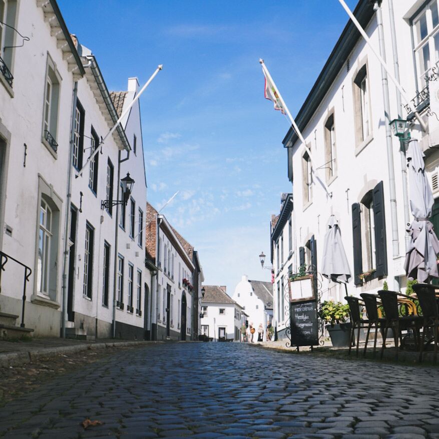 Een smalle kasseienstraat geflankeerd door witte huizen met donkere luiken en vlaggenmasten. Aan de rechterkant staan tafels en stoelen van een terras met opgevouwen parasols. De lucht is helderblauw met enkele lichte wolken.