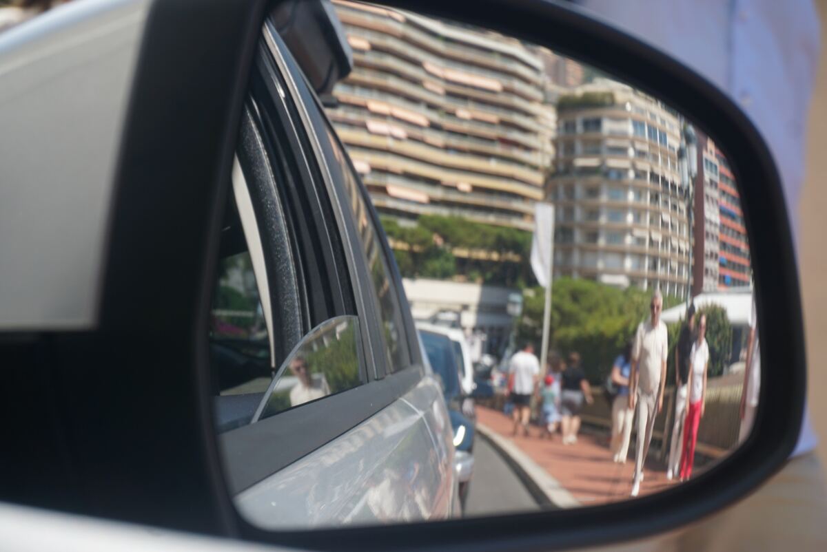 Een close-up vanuit een auto, gericht op de rechter buitenspiegel. In de spiegel is een stadsgezicht te zien met een straat, een roodbruin trottoir en verschillende voetgangers. Op de achtergrond staan hoge gebouwen met balkons en er zijn groene bomen zichtbaar. Buiten de spiegel is een deel van de auto en een wazige reflectie van gebouwen te zien. Het is een zonnige ochtend.