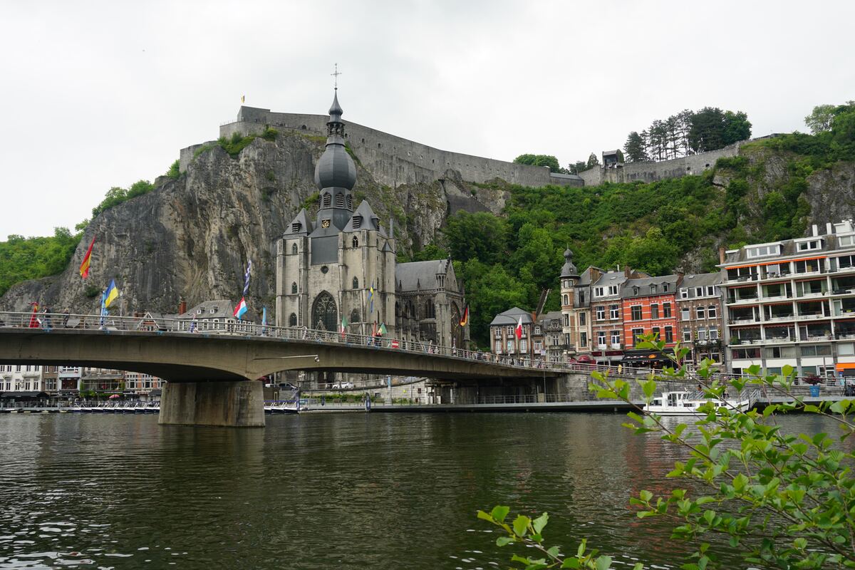 Een brede foto van de stad Dinant met de Maas op de voorgrond, een brug met vlaggen, de Collegiale kerk van Onze-Lieve-Vrouw en de citadel op de rotswand. Aan de rechterkant staan diverse gebouwen langs de rivier.