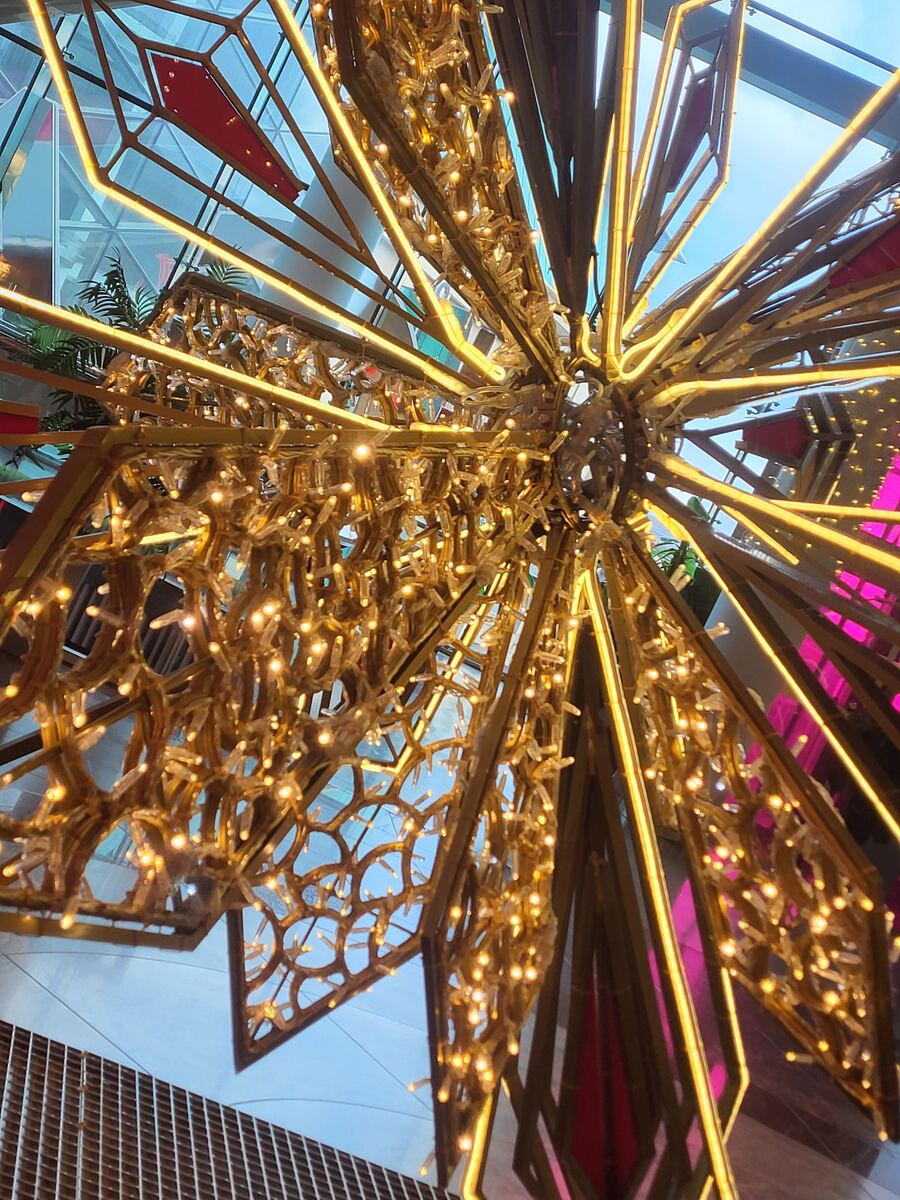 Een close-up van een grote, stervormige decoratie met veel kleine lampjes en gouden accenten. Op de achtergrond is een glazen plafond of dak zichtbaar, met daarachter een blauwe lucht en enkele groene planten.