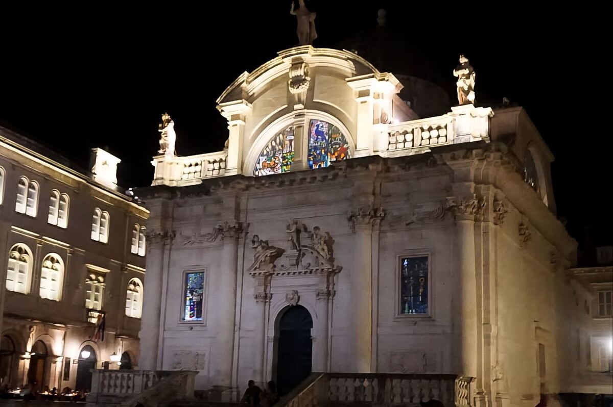 Een foto van een verlichte kerk met een barokke gevel en glas-in-loodramen, 's nachts in Dubrovnik. De kerk heeft beelden op het dak en boven de ingang. Links van de kerk staat een ander gebouw met verlichte ramen. Op de voorgrond zijn mensen te zien die op een trap zitten.