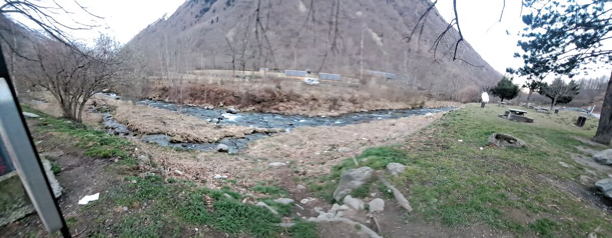 Een panoramische foto van een berglandschap met een rivier die door een vallei stroomt. Op de voorgrond is een groene oever te zien met enkele picknicktafels en bomen. De bergen zijn deels begroeid met kale bomen en struiken, en de lucht is licht bewolkt. Rechts is een deel van een weg zichtbaar.