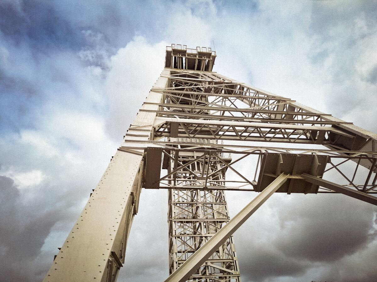 Een laag camerastandpunt toont een grote, metalen schachtbok van een mijn, oprijzend tegen een bewolkte lucht. De constructie bestaat uit een complex netwerk van balken en spanten, met een platform bovenaan.