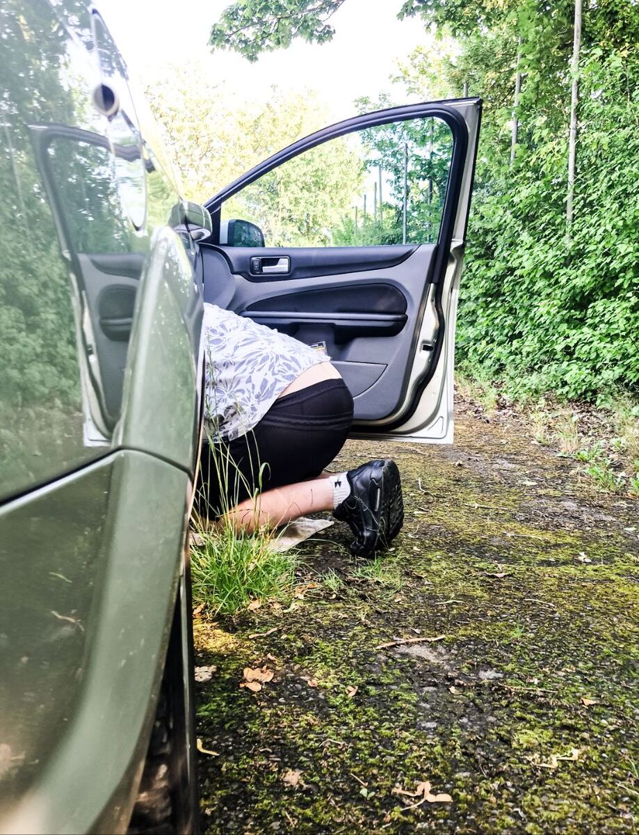 Een persoon in een wit overhemd met print en zwarte korte broek knielt naast een open autodeur van een groene Ford Focus stationwagen. De persoon kijkt onder de auto, die op een onverharde ondergrond met gras en mos staat. Op de achtergrond is groen gebladerte van bomen en struiken te zien.