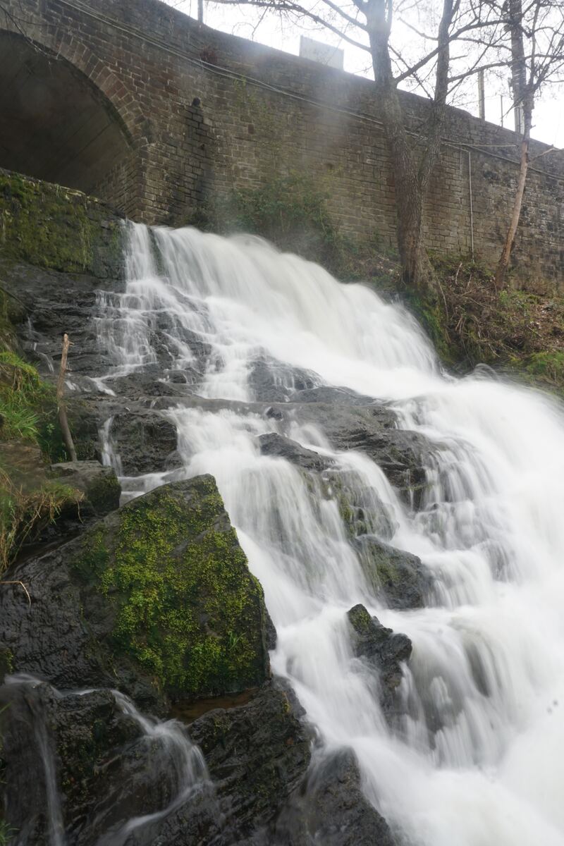 Een waterval stroomt over rotsen en mos naar beneden, met op de achtergrond een stenen brug en een muur. De bomen boven de muur zijn kaal.