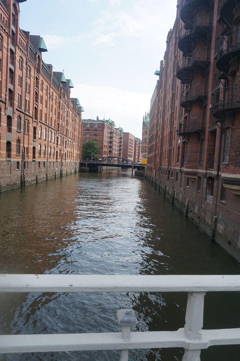 De foto toont een smalle waterweg die wordt omzoomd door hoge, bakstenen pakhuizen aan beide zijden. De gebouwen hebben veel ramen en karakteristieke daken, waarvan sommige met groene koperen accenten. In de verte overspant een brug de waterweg. De lucht is deels bewolkt en het wateroppervlak reflecteert de omliggende architectuur. Een wit hekwerk is op de voorgrond zichtbaar.