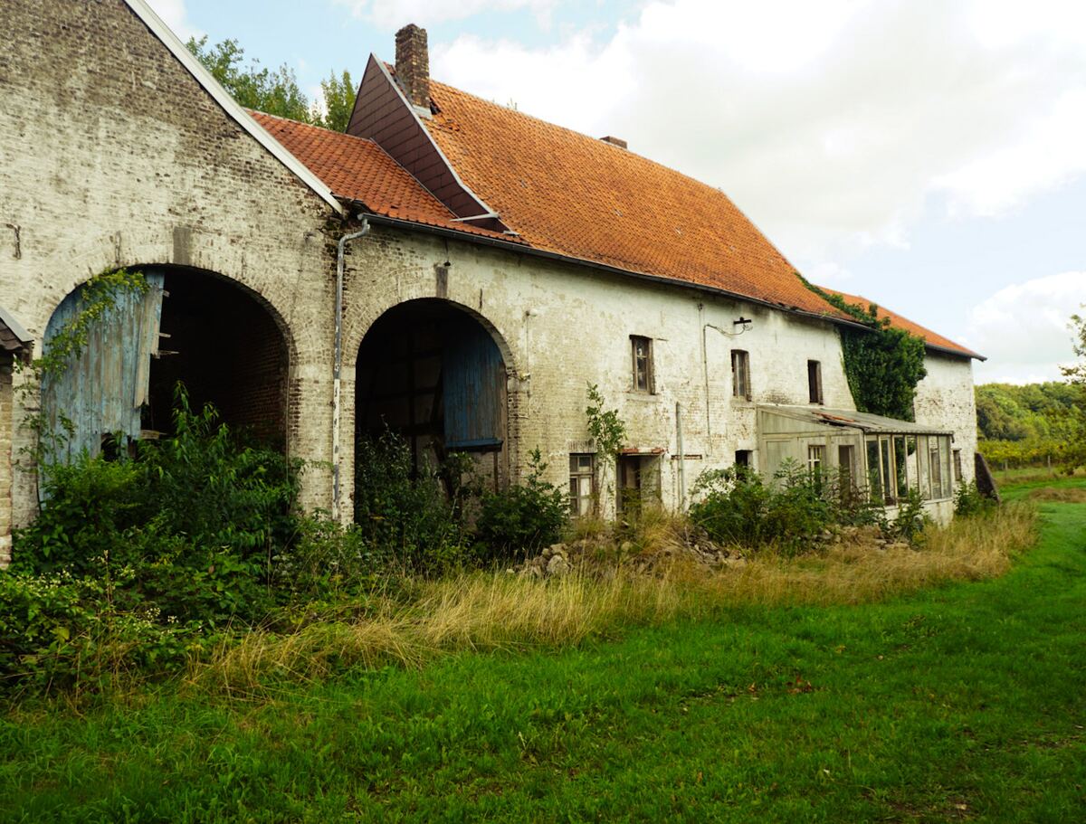 Een oude, deels vervallen boerderij met witte muren en een oranje pannendak, omringd door overwoekerd groen en gras. De boerderij heeft twee grote boogvormige ingangen met deels kapotte blauwe deuren en een aangebouwde serre.