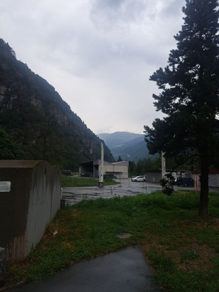 De foto toont een uitzicht op een bergachtig landschap onder een bewolkte hemel. Op de voorgrond links staat een grote, grijze betonnen constructie. Rechtsvoor is een deel van een grote, donkergroene naaldboom zichtbaar. In het midden van de foto bevindt zich een verhard terrein, mogelijk een parkeerplaats of industrieterrein, met daarop een wit voertuig en een donkerder voertuig. Aan de achterzijde van dit terrein staat een gebouw met een donker dak en lichtgrijze muren, dat lijkt op een loods