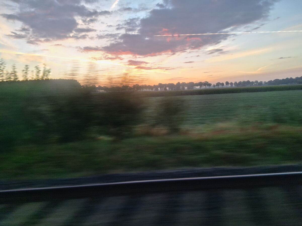 Een foto genomen vanuit een rijdend voertuig toont een landschap met groene velden en een rij bomen aan de horizon. De zon komt op en kleurt de lucht met oranje, roze en paarse tinten. Er zijn enkele wolken en vliegtuigstrepen zichtbaar. Op de voorgrond is een deel van een vangrail of reling te zien, wat de indruk geeft van beweging.