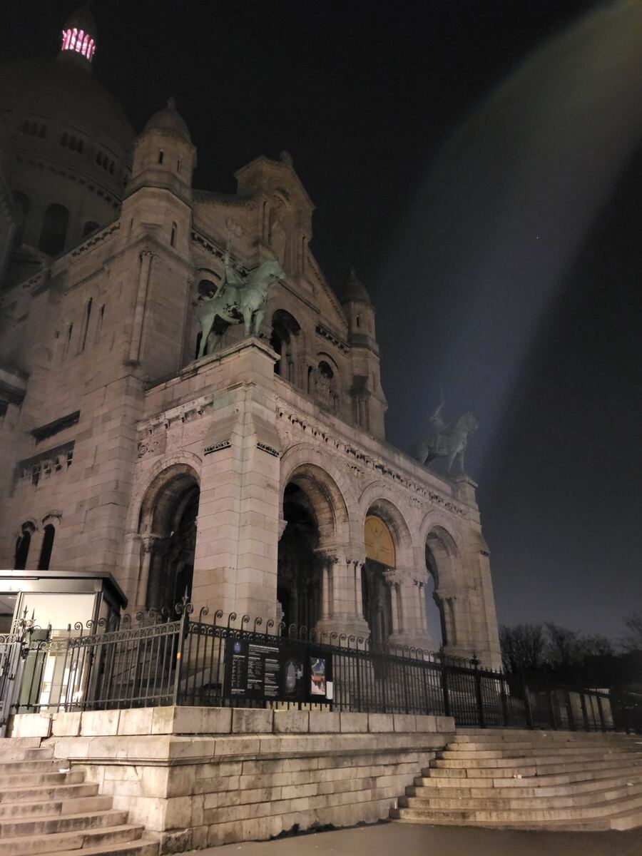 De foto toont de gevel van een grote, lichtgekleurde basiliek in de nacht, met een donkere lucht erboven. Er zijn bogen, pilaren en beelden van ruiters op paarden zichtbaar. Een deel van de koepel met roze verlichting is linksboven te zien. Op de voorgrond zijn stenen trappen en een metalen hekwerk.