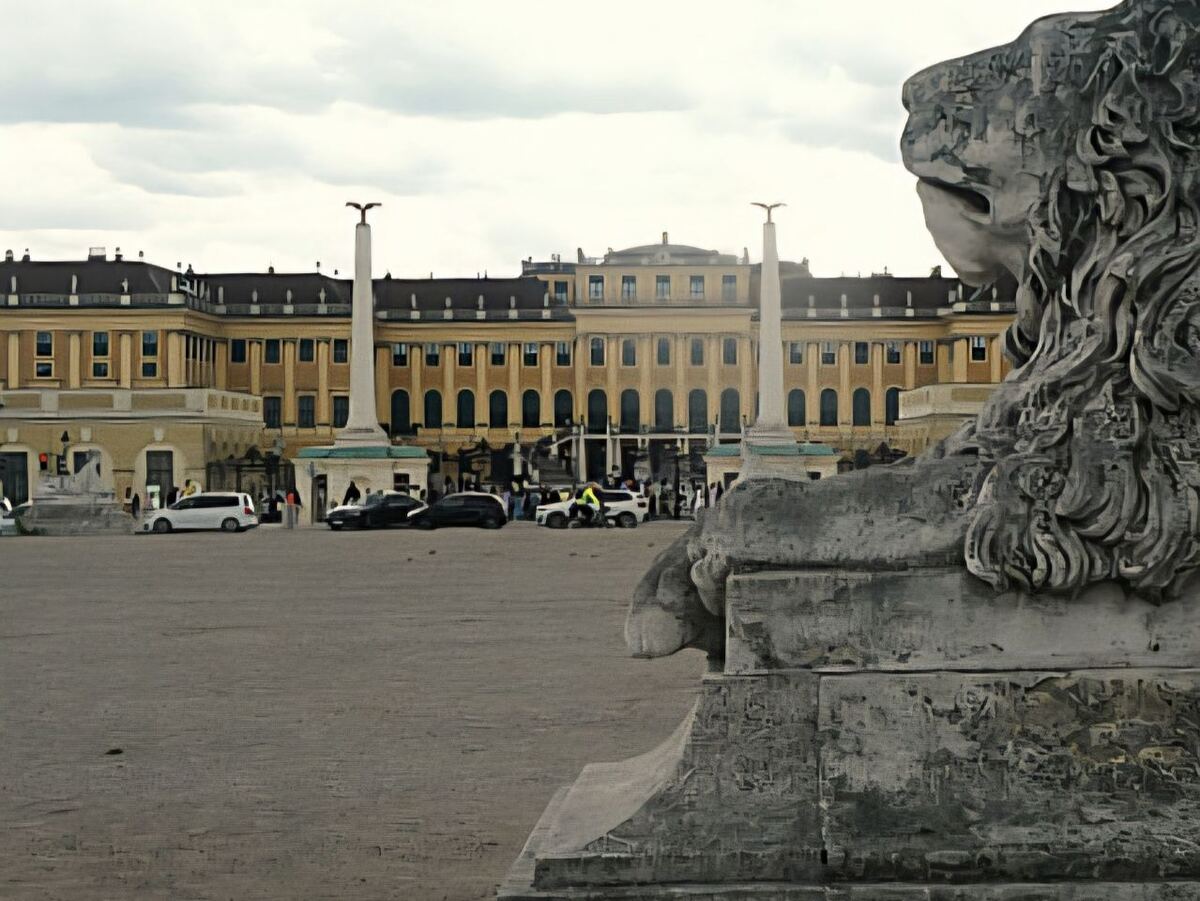Een foto van het paleis Schönbrunn in Wenen, gezien vanaf een plein. Op de voorgrond is een deel van een stenen leeuwenbeeld te zien. Op het plein staan enkele auto's en mensen. De lucht is bewolkt.