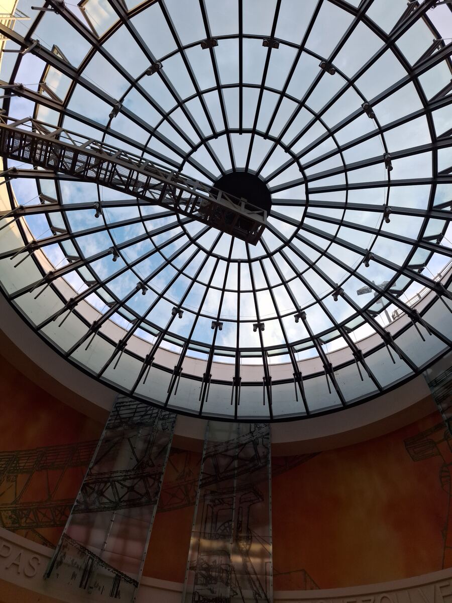 Een foto van een grote glazen koepel, gezien van onderaf. De koepel heeft een metalen rasterstructuur en laat de blauwe lucht met enkele wolken zien. In het midden hangt een metalen constructie, mogelijk voor onderhoud of verlichting. De wanden eronder zijn oranje getint met industriële tekeningen en er hangen glazen panelen met daarop ook industriële afbeeldingen.