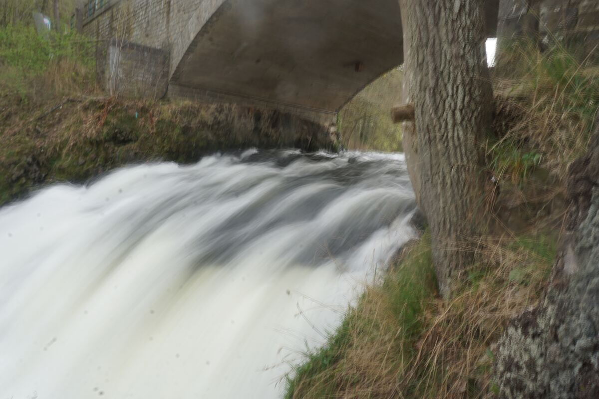 Een close-up van snelstromend water onder een stenen brug, met een boomstam aan de rechterkant en begroeiing op de oevers. Het water is wazig door de lange sluitertijd, wat de beweging benadrukt.