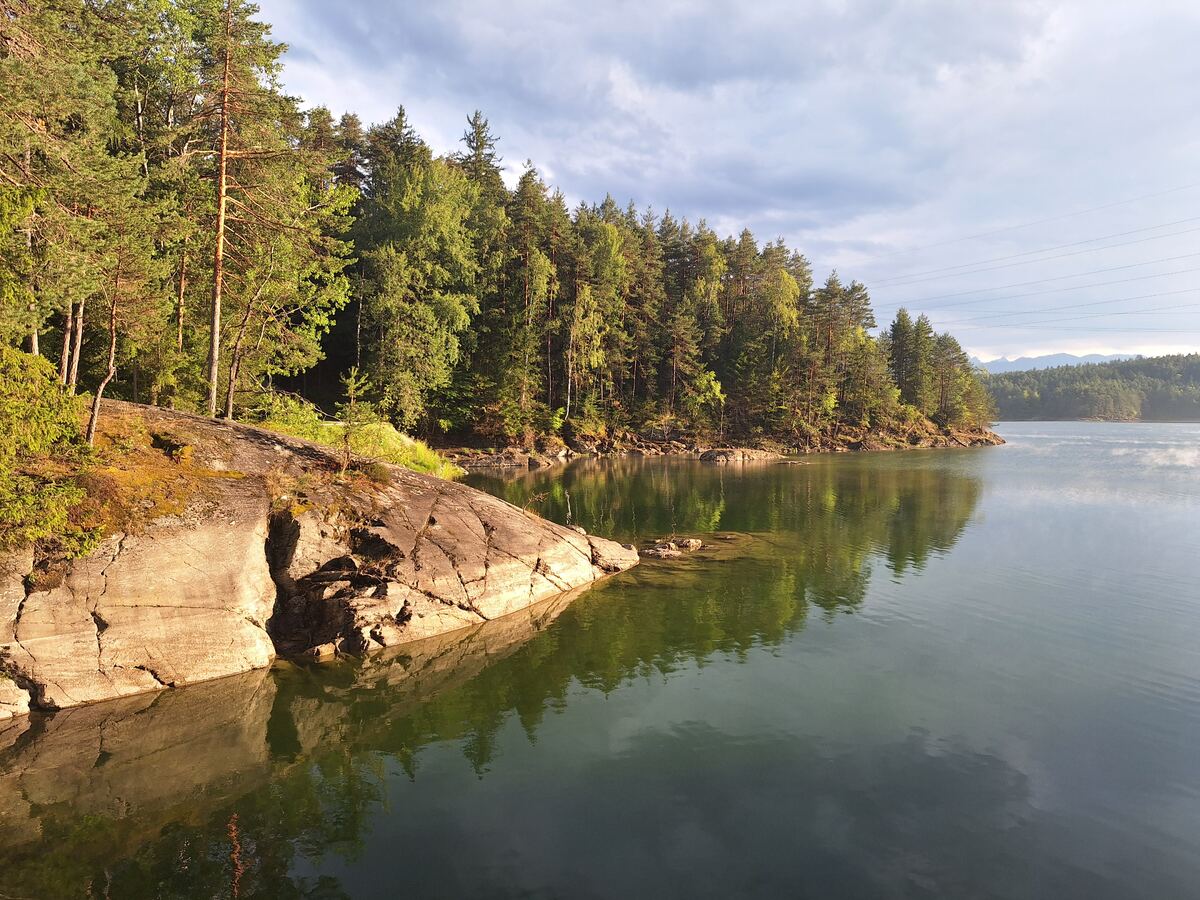 Een rustig meer met aan de linkerkant een rotsachtige oever begroeid met bomen. Het water reflecteert de bomen en de lucht. Aan de horizon zijn bergen zichtbaar onder een bewolkte hemel.