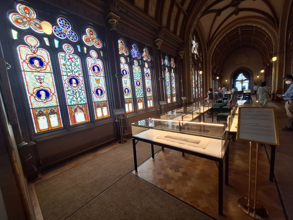 Een lange hal met een reeks hoge, gedetailleerde glas-in-loodramen aan de linkerkant. De ramen tonen portretten en heraldische motieven in levendige kleuren. In het midden van de hal staan glazen vitrines met daarin tentoongestelde voorwerpen. De vloer bestaat deels uit parket en deels uit tapijt. Aan het einde van de hal zijn mensen te zien die rondkijken.