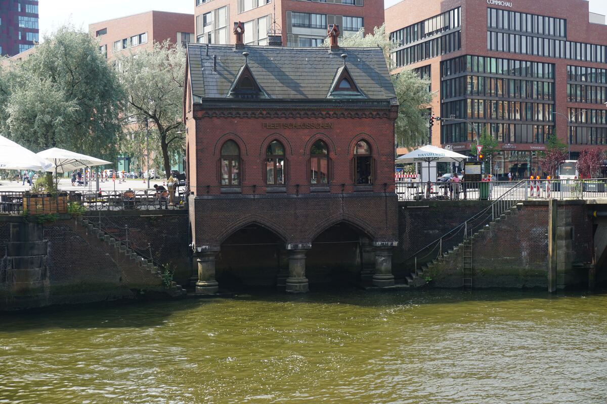 Een bakstenen gebouw met de naam 'FLEETSCHLÖSSCHEN' staat aan het water, ondersteund door bogen en pilaren. Op de achtergrond zijn moderne gebouwen, bomen en een terras met parasols zichtbaar.