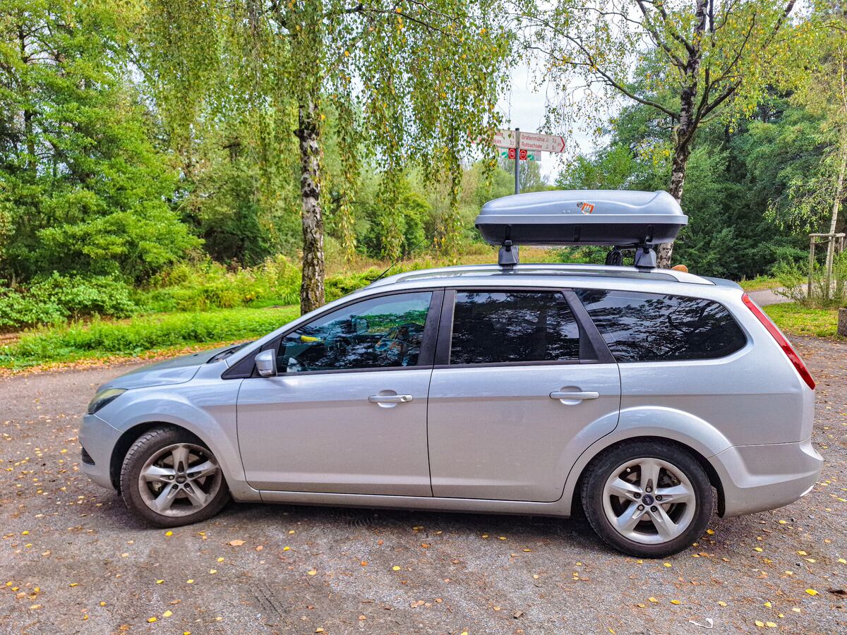 Een zilverkleurige Ford Focus stationwagen met een grijze dakkoffer op het dak staat geparkeerd op een onverhard pad. Op de achtergrond zijn bomen met groen en deels geel blad te zien, en een wegwijzer met Duitse plaatsnamen.