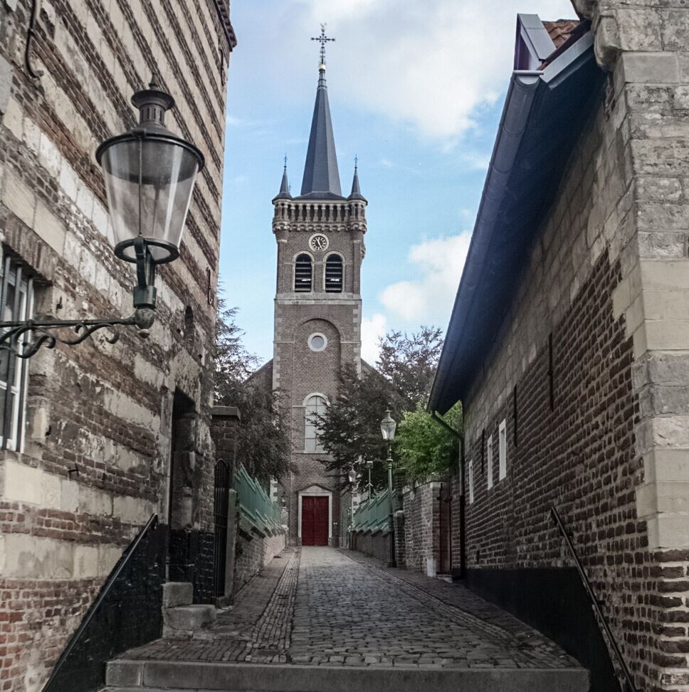 Een smalle, geplaveide steeg leidt naar een kerk met een hoge spits en een rode deur. Aan weerszijden van de steeg staan oude bakstenen gebouwen, waarvan één met een opvallend gestreept patroon en een ouderwetse lantaarn.
