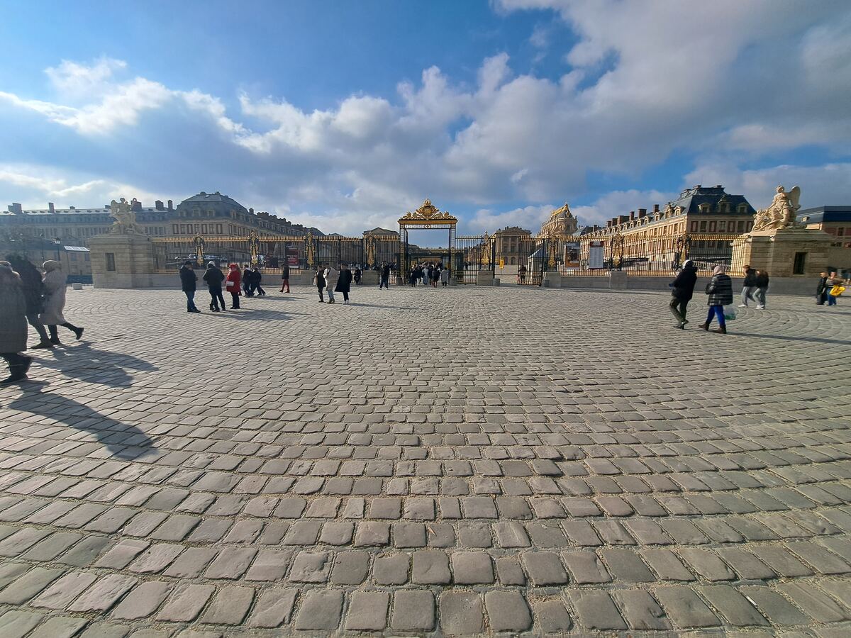 Een weidse blik op een groot, geplaveid plein met op de achtergrond de gouden poorten van een historisch gebouw. Er lopen diverse mensen over het plein en de lucht is deels bewolkt met een helderblauwe hemel.