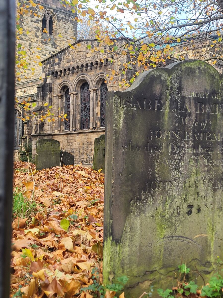 Een oude, stenen kerk met romaanse bogen en gebrandschilderde ramen, deels zichtbaar door takken met herfstbladeren. Op de voorgrond liggen veel bruine en gele bladeren op de grond, waartussen meerdere oude, met mos begroeide grafstenen staan. Een van de grafstenen toont duidelijk leesbare inscripties.