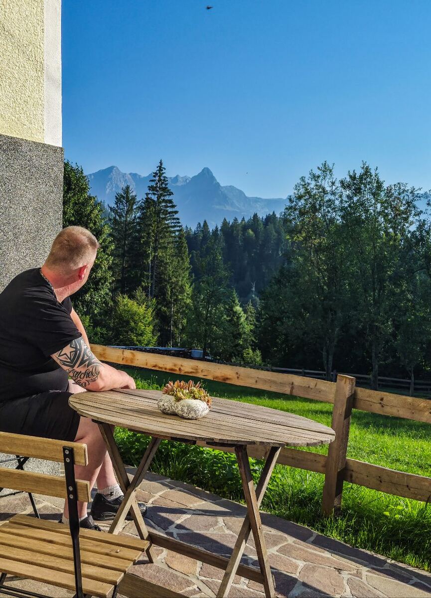 Een man zit aan een houten tafel op een terras en kijkt uit over een groen landschap met bossen en bergen op de achtergrond. De lucht is helderblauw. Op de tafel staat een plantje in een stenen pot.