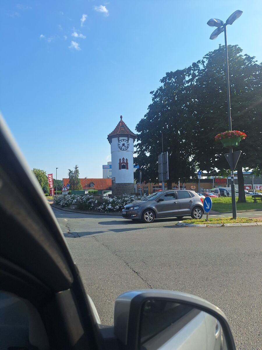 De foto is genomen vanuit een auto en toont een witte toren met een rode puntdak en een klok aan een rotonde. Rondom de toren staan witte bloeiende struiken. Op de achtergrond is een grote groene boom zichtbaar onder een helderblauwe lucht. Een grijze auto rijdt op de rotonde. In de voorgrond zijn delen van een auto en een zijspiegel zichtbaar.