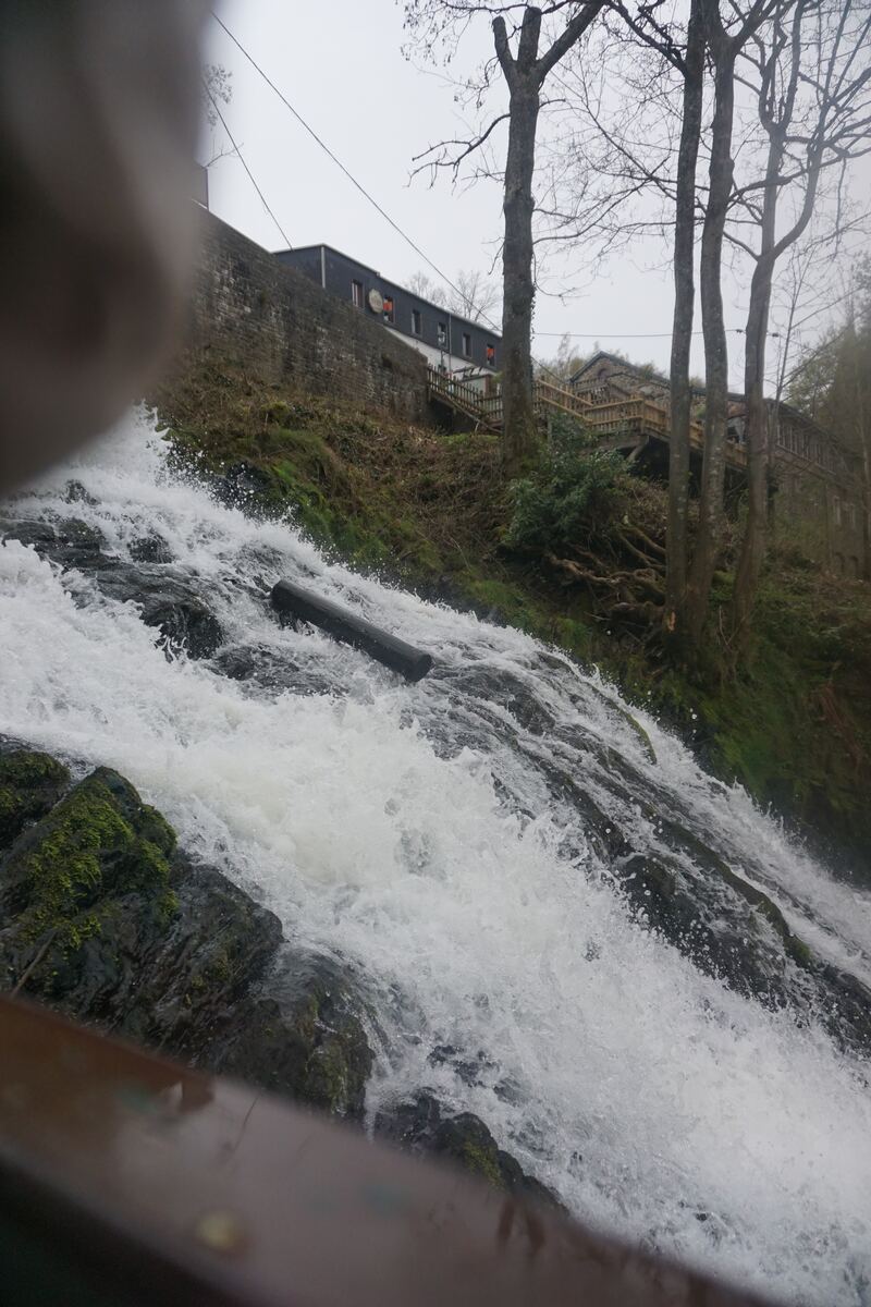 Een foto van een waterval die over rotsen stroomt, met op de achtergrond een heuvel met bomen en een gebouw. Een boomstam ligt in het water van de waterval. De lucht is grijs en bewolkt.
