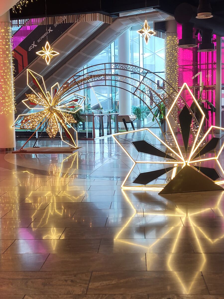 De foto toont een interieur van een modern gebouw, waarschijnlijk een winkelcentrum of bioscoop, met feestelijke verlichting. Grote, stervormige lichtobjecten staan op de glimmende vloer en worden weerspiegeld. Op de achtergrond zijn roltrappen en grote ramen te zien, met een neonroze gloed aan de rechterkant en een bord met '4DX SCREEN X'.