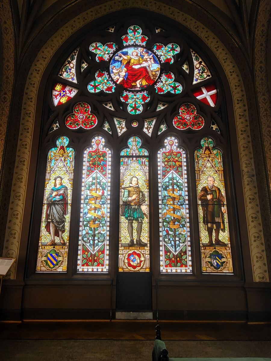 Een groot, gedetailleerd glas-in-loodraam met een centrale ronde afbeelding van een figuur met engelen, omringd door kleinere decoratieve panelen en vier verticale panelen met afbeeldingen van historische figuren. De kleuren zijn levendig en de details van de figuren en patronen zijn duidelijk zichtbaar. De architectuur rondom het raam is versierd met patronen.