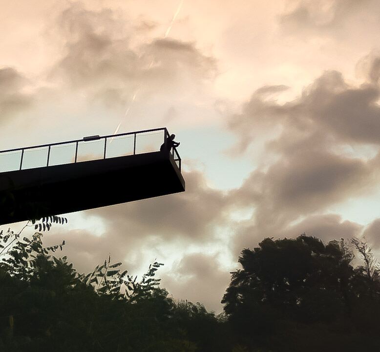 Een silhouet van een persoon staat op een modern uitzichtplatform met glazen balustrade, uitkijkend over een bosrijk landschap. De lucht is bewolkt met een lichte oranje gloed van de ondergaande zon.