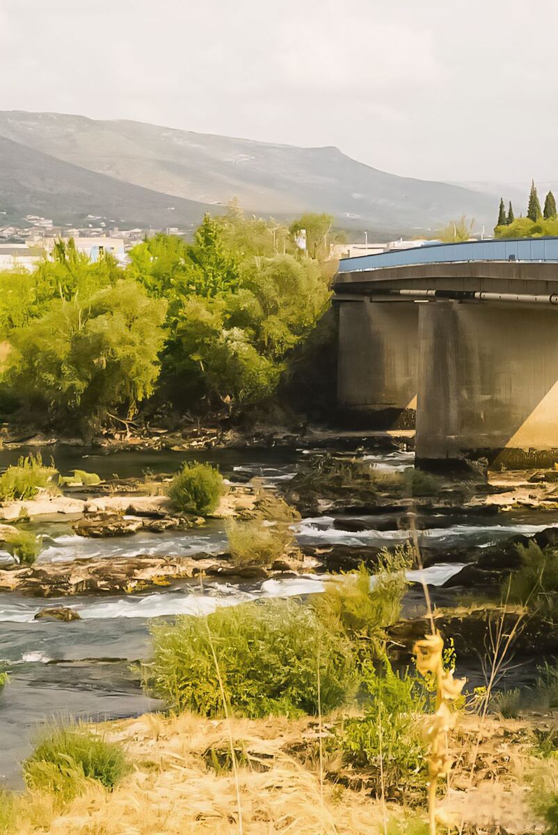 Een verticale foto van de Neretva rivier met een moderne brug op de achtergrond. De rivier stroomt over rotsen en door begroeiing, met bergen en bebouwing in de verte onder een lichte hemel.