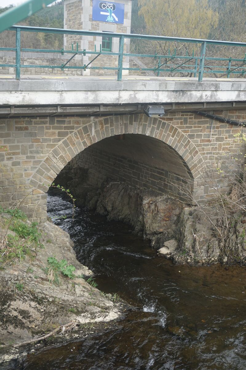 Een stenen boogbrug over een beek met rotsachtige oevers. Bovenop de brug is een groen metalen hekwerk zichtbaar en een blauw bord met 'Cascade Coo'. Op de achtergrond zijn bomen en een heuvel te zien.