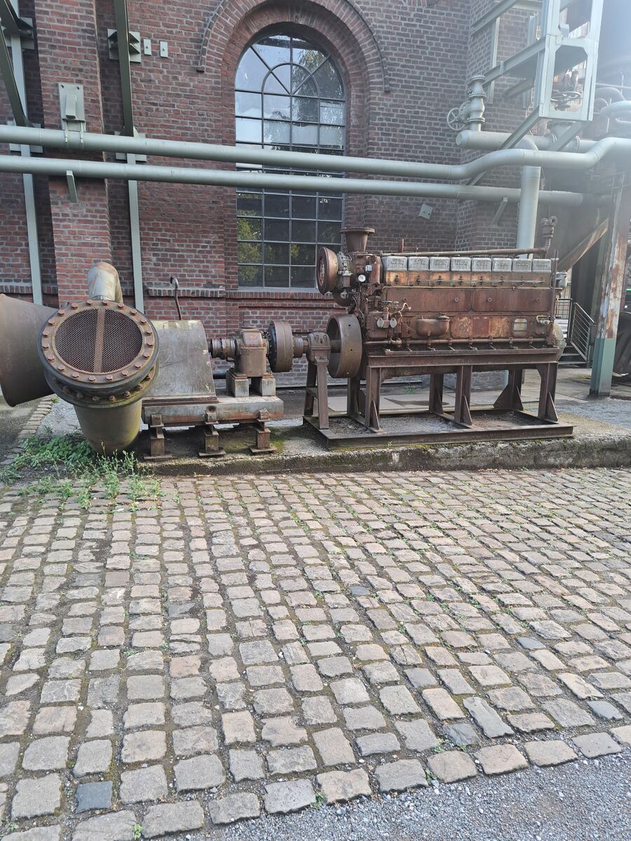 Een roestige, grote industriële machine, mogelijk een motor, met een bijbehorende pijp of ventilator staat op een geplaveid oppervlak van kasseien. Op de achtergrond is een bakstenen gebouw te zien met grote, ronde boogvensters en diverse leidingen langs de muur. Kleine plantjes groeien tussen de kasseien.