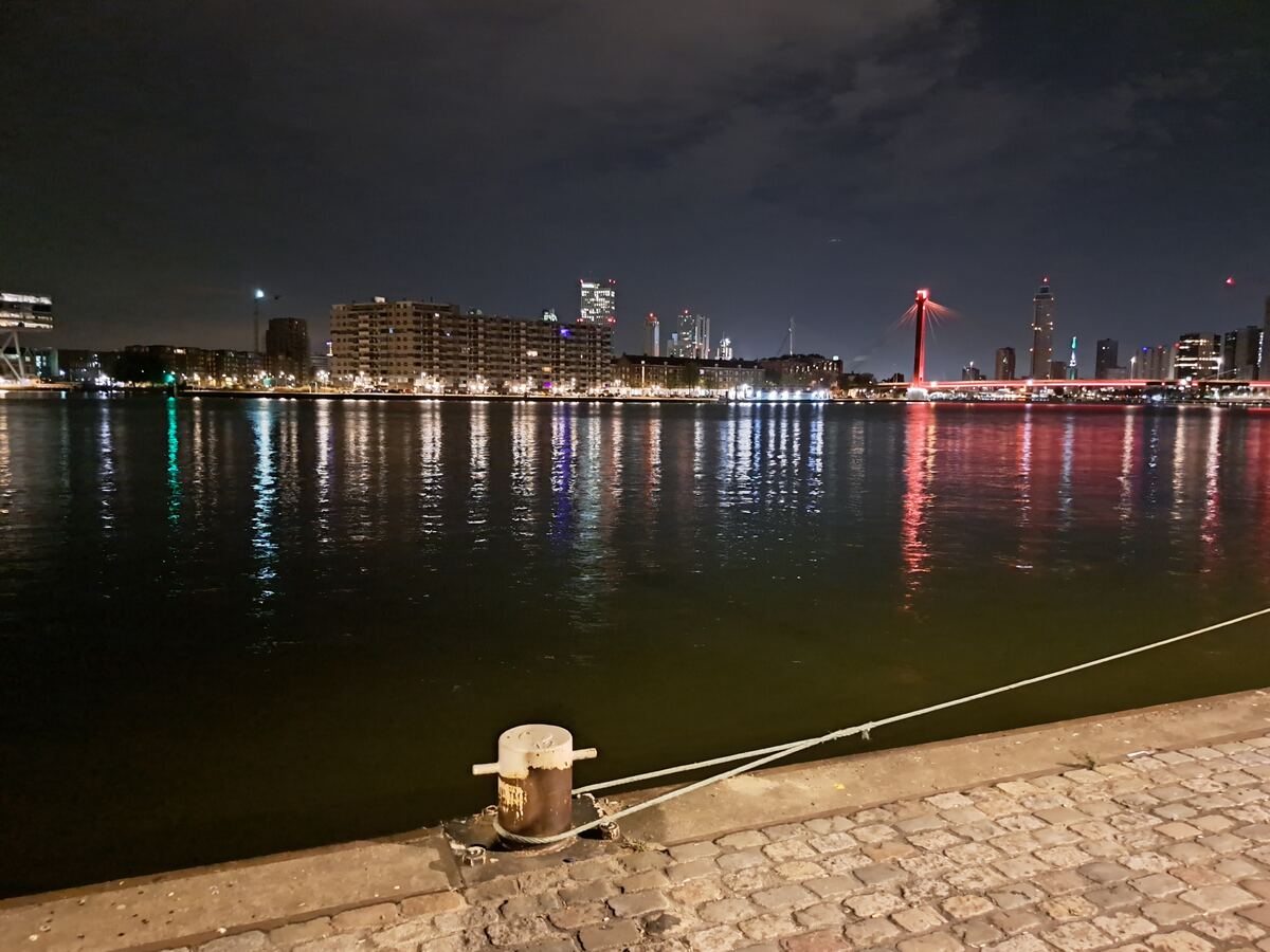 Een nachtelijke opname van de Maas in Rotterdam, met de skyline van de stad en de Erasmusbrug verlicht op de achtergrond. Op de voorgrond is een deel van de kade met kasseien en een bolder te zien, waaraan een touw is bevestigd. De lichten van de gebouwen en de brug reflecteren in het water.