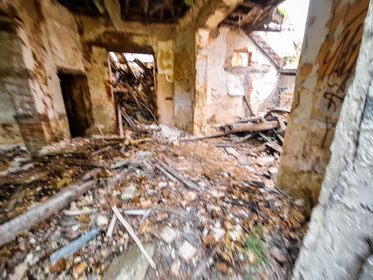 De foto toont het interieur van een vervallen gebouw, met puin, houten balken en brokstukken verspreid over de grond. Muren zijn deels ingestort en er zijn openingen waar ramen of deuren moeten zijn geweest, met uitzicht op wat groen buiten. De algehele indruk is van verwaarlozing en verval.