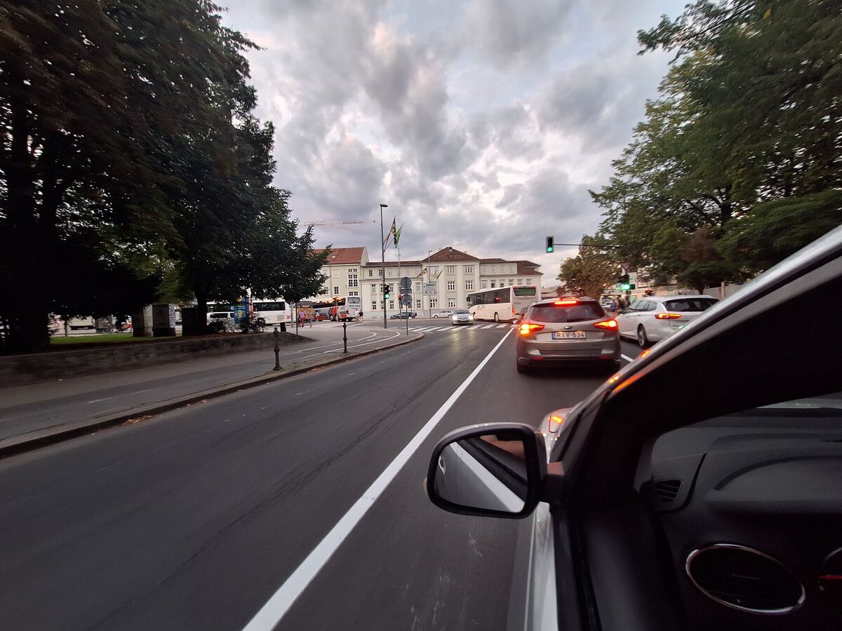 We kijken vanuit de auto naar een drukke straat in een stad, met meerdere auto's en bussen op de weg. Aan de linkerkant van de weg staan bomen en een groot gebouw met meerdere verdiepingen en ramen. De lucht is bewolkt en de verkeerslichten zijn groen.