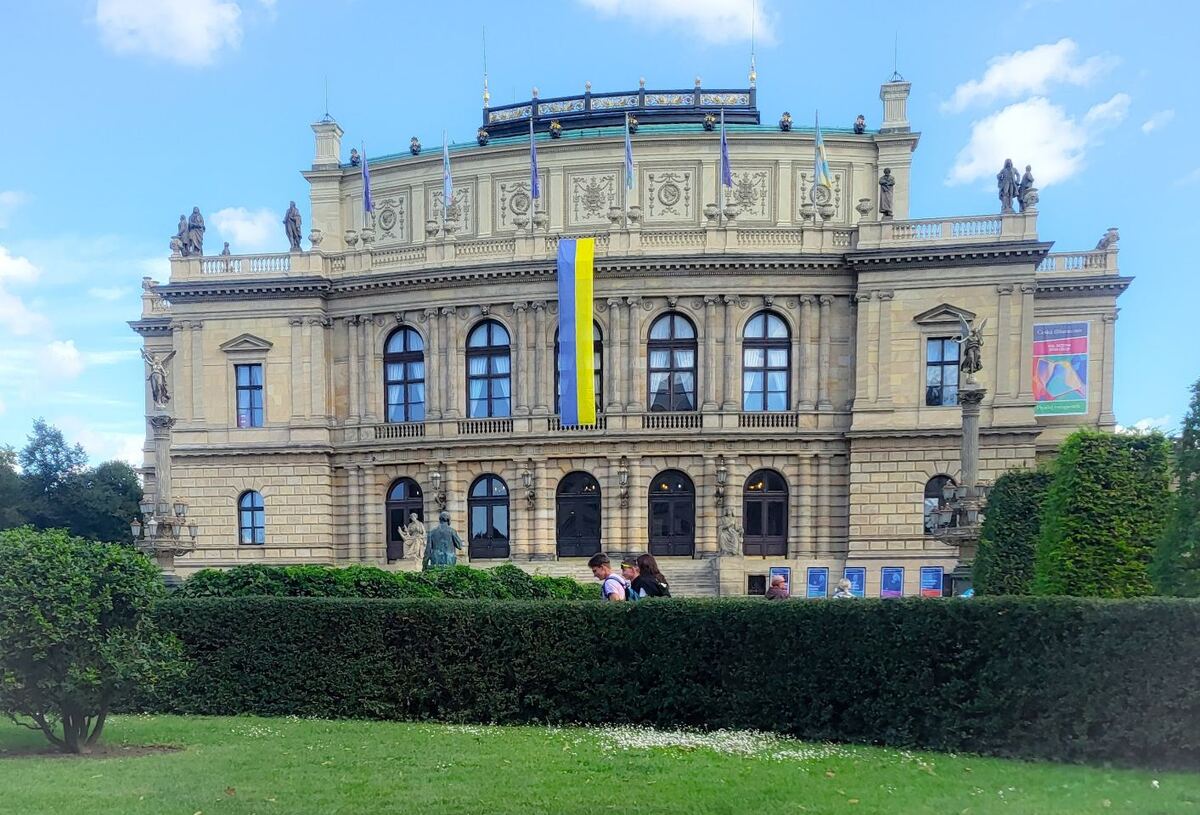 Een breed shot van het Rudolfinum in Praag, een neorenaissancegebouw met een vlag van Oekraïne aan de gevel. Op de voorgrond is een groene haag en een grasveld te zien, met enkele mensen die achter de haag lopen.