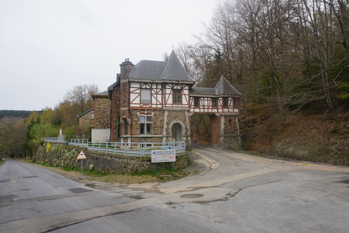 Een stenen gebouw met vakwerk en een overspanning over een weg, gelegen aan een splitsing in een bosrijke omgeving. Een verkeersbord voor een gevaarlijke daling is zichtbaar, evenals een bord met 'CENTRE HOSPITALIER PELTZER-LA TOURELLE'.