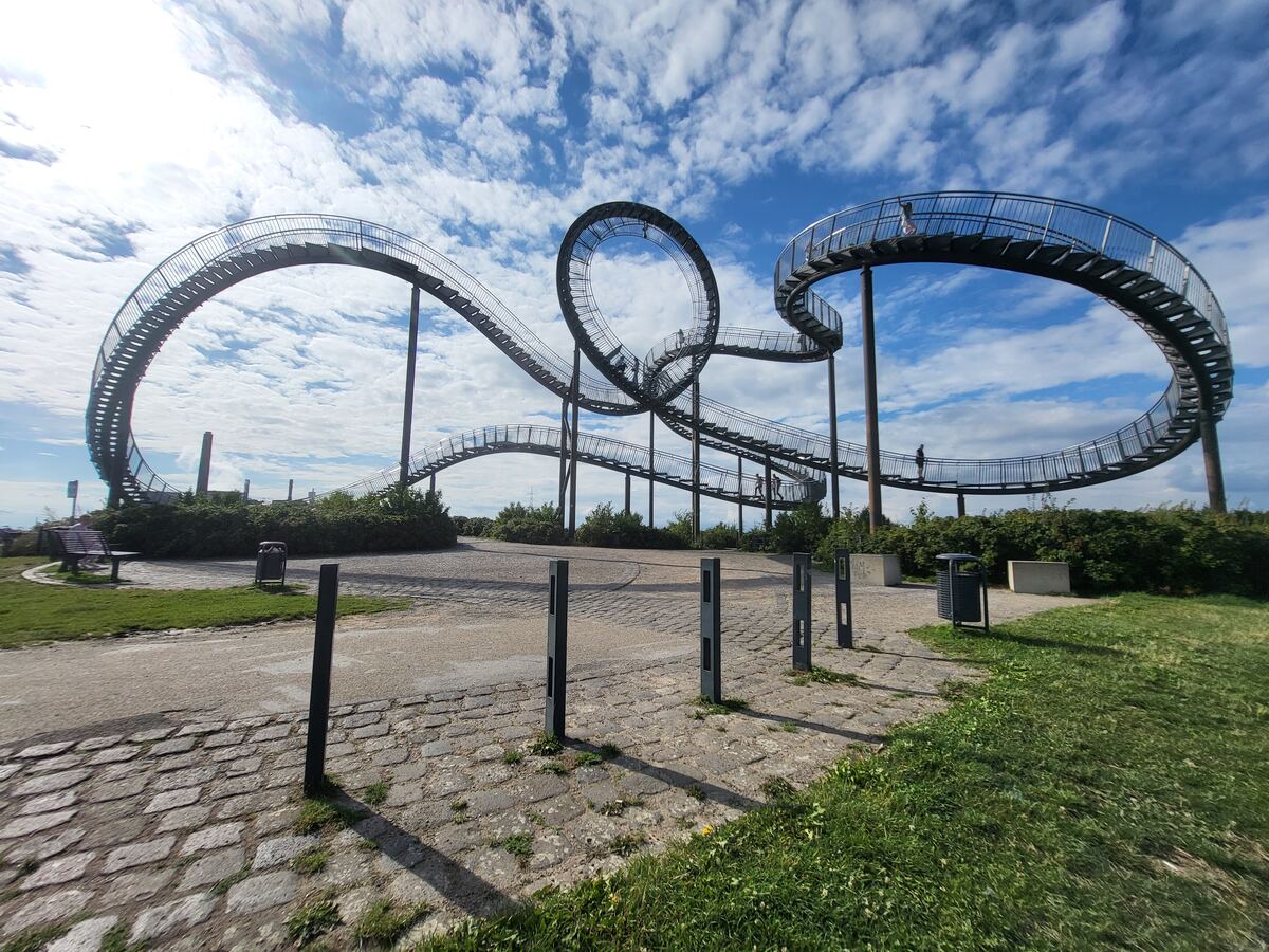 Een brede opname van de Tiger & Turtle – Magic Mountain, een sculptuur in de vorm van een wandelbare achtbaan, met mensen die over de paden lopen. De sculptuur is gemaakt van metaal en staat op een heuvel met groene begroeiing. De lucht is blauw met witte wolken.