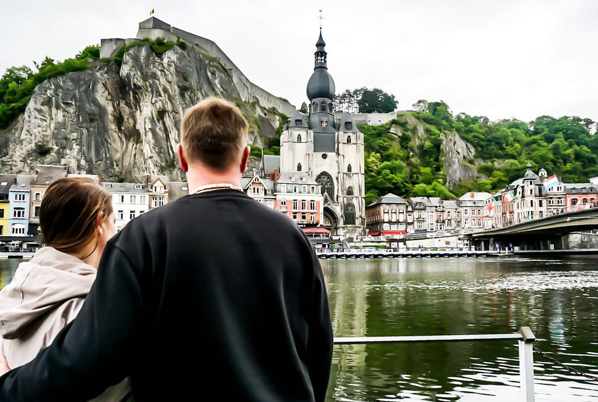 Een stel kijkt uit over de Maas in Dinant, met op de achtergrond de Citadel van Dinant en de Collegiale kerk van Onze-Lieve-Vrouw. De rivier is rustig en weerspiegelt de lucht. Aan de overkant van het water staan kleurrijke gebouwen en een brug.