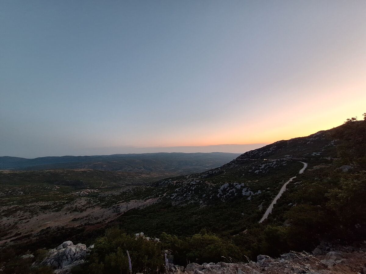Een weids landschap met bergen en een kronkelende weg, gefotografeerd tijdens zonsondergang. De lucht vertoont een zachte overgang van blauw naar oranje. De voorgrond bestaat uit rotsachtig terrein met lage begroeiing, terwijl in de verte heuvels en dalen zichtbaar zijn.