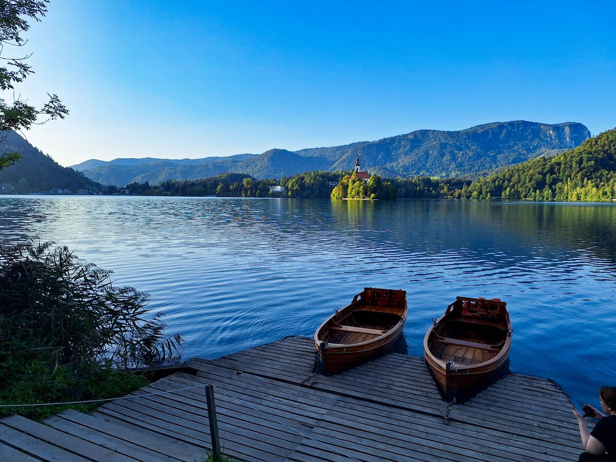 Een houten steiger met twee roeiboten op het water van een meer. Op de achtergrond is een eiland met een kerk te zien, omringd door beboste heuvels en bergen onder een helderblauwe lucht. Rechtsonder is een persoon de foto aan het maken.