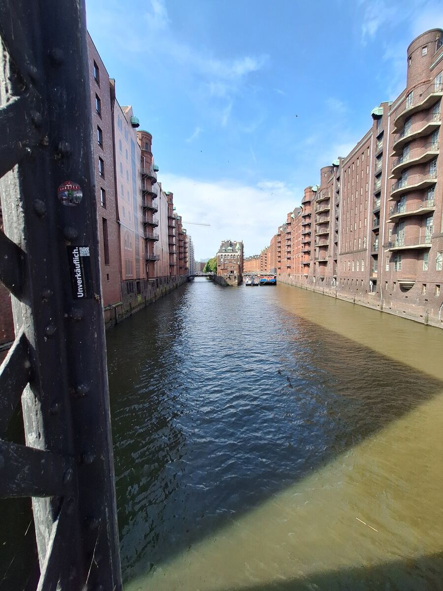 Een foto genomen vanaf een brug, kijkend over een kanaal met aan weerszijden hoge bakstenen pakhuizen. De lucht is blauw met enkele wolken en de zon schijnt, wat zorgt voor een schaduw van de brug op het water. Aan het einde van het kanaal is een ander gebouw zichtbaar en liggen er enkele boten.