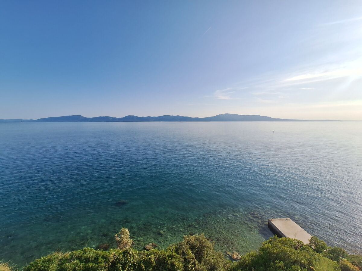 Een breed panoramisch uitzicht over de Adriatische Zee met op de achtergrond een rij bergen. Op de voorgrond is een groene kustlijn te zien met een kleine betonnen steiger die in het heldere water steekt. De lucht is blauw met enkele lichte wolken en de zon schijnt vanaf rechtsboven.